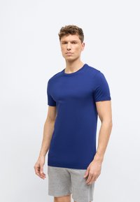 Νεαρός άνδρας με κοντά σγουρά μαλλιά, φορώντας εφαρμοστό σκούρο μπλε μπλουζάκι και ανοιχτό γκρι σορτς, στέκεται μπροστά σε απλό λευκό φόντο.