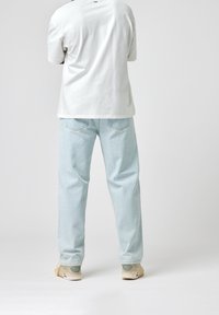 Biała koszulka z krótkim rękawem, jasnoniebieskie dżinsy oraz beżowe trampki. Prosty design, swobodny fason, widoczne szwy i subtelne logo z tyłu koszulki.