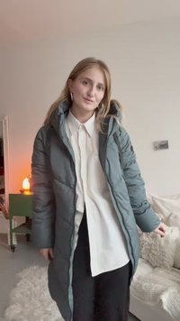 Pufīgs garš mētelis tumši zilā krāsā ar kapuci, ar rāvējslēdzēja priekšu, sānu kabatām un teksturētu audumu, uzvilkts virs baltas krekla.