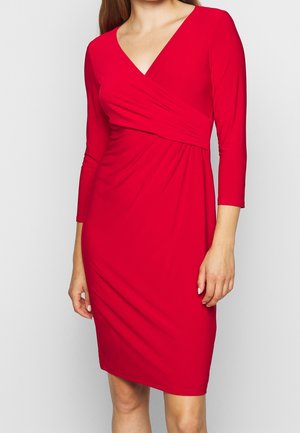 Femme portant une robe rouge mi-longue aux genoux avec des manches trois-quarts et un décolleté en V portefeuille, debout devant un fond clair uni.