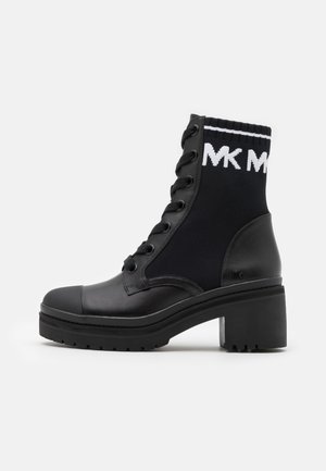 Czarny skórzany i dzianinowy botek za kostkę z przodu na sznurowadła, z grubym obcasem i białą opaską z logo "MK" na górnym mankiecie na białym tle.
