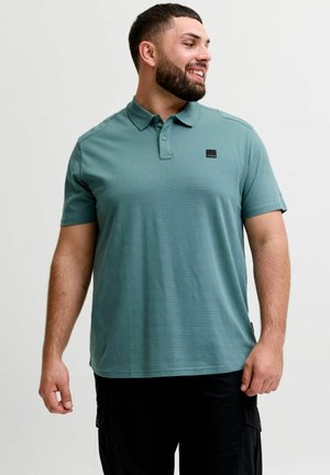 Rövid sötét hajjal és szakállal rendelkező férfi mosolyog, türkizkék rövid ujjú pólót és fekete nadrágot visel, egy egyszínű háttér előtt áll.