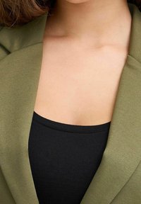 Blazer vert à finition texturée, associé à un débardeur noir ajusté. L'image met en valeur le décolleté et la zone des épaules.