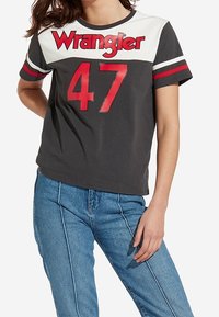 Šedé tričko s krátkým rukávem s nápisem "Wrangler" červeným písmem a číslem "47" v kontrastní červené barvě, s bílými akcenty a červenými pruhy na rukávech.