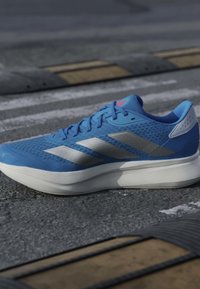 Basket athlétique bleue avec semelle blanche et bandes argentées posée sur un bitume texturé près d’un ralentisseur noir et jaune.