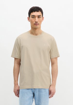 Homme debout devant un fond blanc, portant un T-shirt beige à manches courtes et un jean bleu clair, faisant face à l'avant avec une expression neutre.