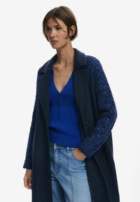 Cappotto navy con maniche a maglia blu strutturata, abbinato a una maglietta blu con bottoni a coste e jeans chiari a vita alta.