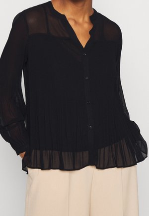 Blouse noire transparente avec boutons, détails plissés et manches longues portée avec un pantalon beige clair.