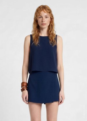 Donna con capelli rossi mossi indossa un top blu navy senza maniche e gonna abbinata, con diversi braccialetti marroni al polso sinistro.