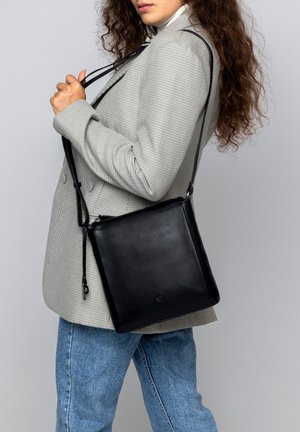 Woman wearing gray checked blazer and blue jeans, holding black leather shoulder bag with long strap against gray background.