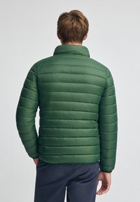 Grön puffjacka med horisontell quiltad design, hög krage och en slät textur. Passformade ärmar och bakvy visas.