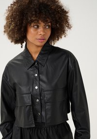 Melnā īsā ādas jaka ar norādītu apkakli, priekšpuses poga un divi krūšu kabatas; gluda tekstūra ar nelielu spīdumu.