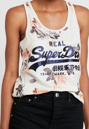 Kobieta ubrana w białą, kwiecistą bluzkę bez rękawów z napisem "Real SuperDry" oraz czarne spodnie, przycięte w talii.