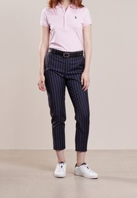 Ljusrosa polo-shirt med en liten logotyp, svart bälte, mörka randiga byxor och vita sneakers med svarta accenter.