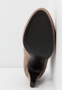 Zapato de tacón alto de ante marrón con punta afilada, que presenta una suela de goma negra texturizada y un diseño de tacón acentuado.
