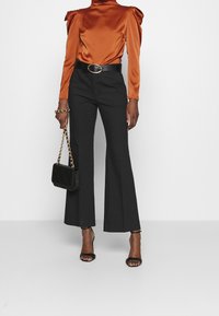 Orangenes Satin-Bluse mit langen Ärmeln und gepufften Schultern, kombiniert mit schwarzen Schlaghosen. Schwarzer Gürtel mit goldenen Beschlägen, passende Handtasche.
