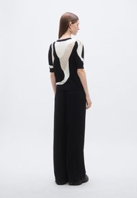 Čierny pletený top s abstraktnými béžovými vzormi, krátkymi rukávmi a priliehavým strihom, v kombinácii s voľnými čiernymi nohavicami a masívnymi topánkami.