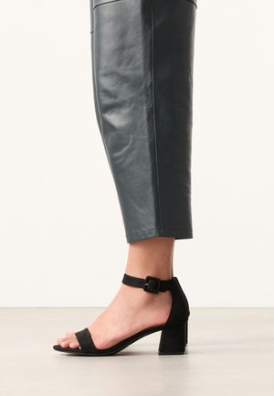Persoon draagt korte donkere leren broek en zwarte sandalen met open tenen, enkelbandje en blokhak, staand op een lichte vloer.