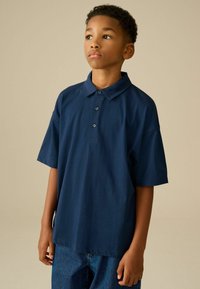 Námořnická modrá polokošile s krátkým rukávem, která má knoflíkovou légu, měkkou látku, uvolněný střih a jednobarevný design, zkombinovaná s denimovými kalhotami.