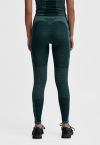 Gröna texturerade leggings med kontrasterande paneler, meshdetaljer och en figurnära design. Kombinerade med svarta träningsskor, sedd bakifrån.