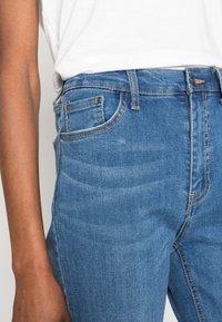 Džíny s vysokým pasem, modré denimové, které mají pět kapes, kontrastní šití a fitted design. Materiál má mírně elastickou strukturu.