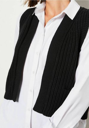 Black knitted vest with ribbed texture, open front design, worn over a white collared shirt. Features short sleeves and a fitted silhouette.