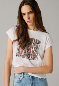 Junge Frau mit langen braunen Haaren, die ein weißes T-Shirt mit dem Aufdruck "PEPE JEANS" und hellblaue Jeans trägt, vor einem grauen Hintergrund stehend.