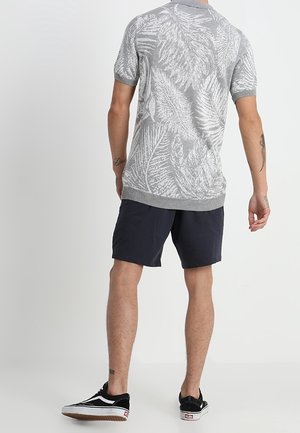 Man draagt een grijs T-shirt met korte mouwen met bladpatroon, marineblauwe korte broek en zwarte sneakers, staand met zijn rug naar de camera op een effen achtergrond.