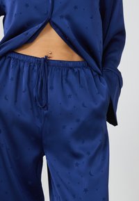 Completo da pigiama blu navy con finitura in raso. Presenta un motivo a stelle e luna, cintura elastica e dettaglio con cordoncino sui pantaloni.