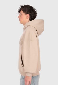 Jeune homme avec des cheveux bouclés et foncés portant un sweat à capuche beige et un jean bleu clair, se tenant de profil sur un fond uni.