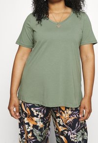 Oliivinvihreä V-kauluksinen ja lyhythihainen t-paita, yhdistetty tummasävyisiin, oransseilla ja valkoisilla yksityiskohdilla koristeltuihin kukkakuosisiin housuihin.