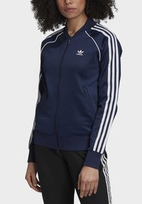 Žena oblečená v tmavě modré adidasácké bundě s bílými pruhy na rukávech a černých kalhotách s bílými pruhy po stranách, stojící na bílém pozadí.