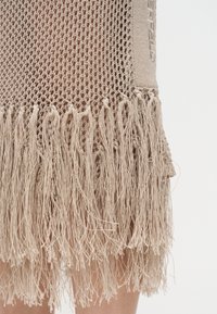 Μπεζ πλεκτό ύφασμα με ανοιχτό πλέγμα και μακριές φούντες που κρέμονται πάνω σε γυμνό δέρμα μπροστά σε λευκό φόντο.