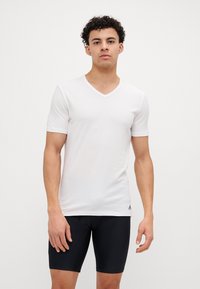 T-shirt blanc à manches courtes et col en V en tissu l�éger et lisse, avec un détail de logo subtil près de l'ourlet. Un short noir ajusté est porté en dessous.