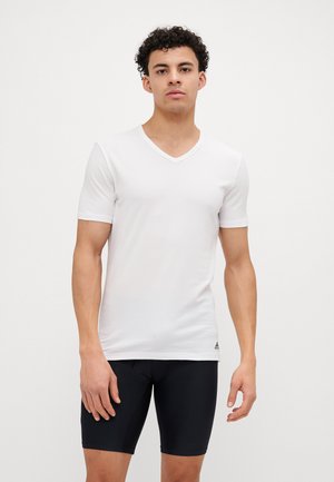 T-shirt blanc à manches courtes et col en V en tissu léger et lisse, avec un détail de logo subtil près de l'ourlet. Un short noir ajusté est porté en dessous.