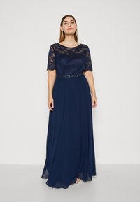 Rochie de seară bleumarin cu mâneci scurte din dantelă și un brâu decorat cu mărgele. Fusta fluidă este realizată dintr-un material moale și ușor.