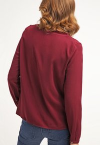 Burgunderfarbene Bluse mit langen Ärmeln aus glattem, leicht glänzendem Material, ausgestattet mit einem schlichten Rundhalsausschnitt und knöpfbaren Manschetten.