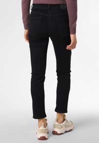 Osoba ve štíhlých černých kalhotách, světle béžových a bílých teniskách a dlouhém mauve svetru se stojí zády k fotoaparátu.