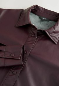 Camisa de piel sintética burdeos con un collar estructurado, cierre de botones y mangas largas con puños redondeados. Textura de forro interior suave.