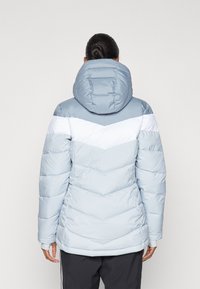 Giacca a vento azzurra chiara con una striscia a zig-zag bianca, dotata di cappuccio e texture trapuntata. La vista posteriore mostra una silhouette aderente e polsini elasticizzati.