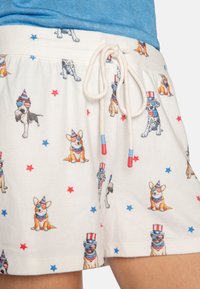 Valkoiset shortsit, joissa on kiristysnauha ja joissa on kuvioita sarjakuvakoirista juhlahatuissa sekä punaisista ja sinisistä tähdistä. Pehmeä materiaali ja rento istuvuus.
