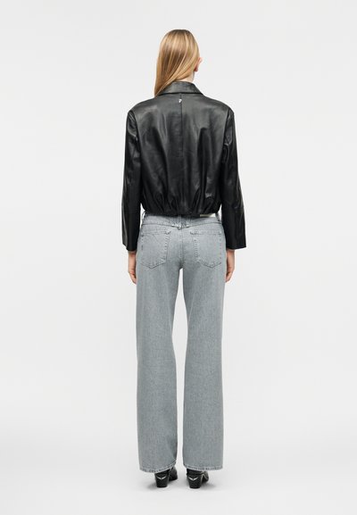Γυναίκα με ίσια ξανθά μαλλιά που φοράει μαύρο δερμάτινο μπουφάν, ανοιχτό γκρι τζιν με φαρδιά μπατζάκια και μαύρα παπούτσια με τακούνι, στέκεται γυρισμένη προς τα πίσω.