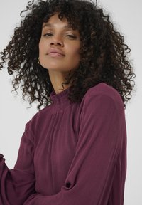 Long-sleeve blouse in deep burgundy, featuring a high smocked neckline and pleated design. Soft texture with a relaxed fit.