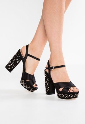 Czarne sandały na wysokim obcasie z satynowego materiału, zaprojektowane w krzyżowy wzór, z teksturowaną platformą ozdobioną złotymi ćwiekami.