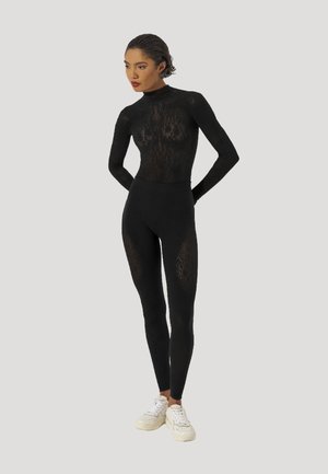 Vrouw staand met handen achter de rug, draagt een zwarte doorschijnende bodysuit met patroon en witte sneakers tegen een effen achtergrond.
