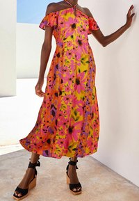 Robe maxi à imprimé floral présentant des couleurs vives de jaune, rose et violet, avec des manches hors épaule et une silhouette fluide, assortie de sandales compensées noires.