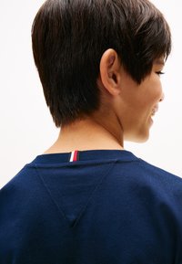 Person with short dark hair facing sideways, wearing a navy blue shirt with a small red, white, and blue striped tag at the neckline.