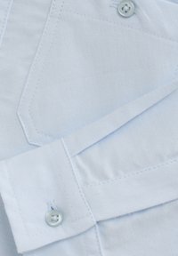 Close-up van lichtblauwe shirtstof, met een soepele textuur, knoopdetail en een puntige zakontwerp met contrasterende stiksels.