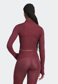 Kvinna med flätat hår i en burgunderröd, långärmad åtsittande topp och matchande högmidjade träningsleggings, sedd från ryggen.