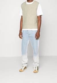 Gilet verde a maglia con coste verticali sopra una maglietta bianca. Jeans azzurri chiari con effetto ombre. Sneaker color sabbia e verde con suola in gomma.
